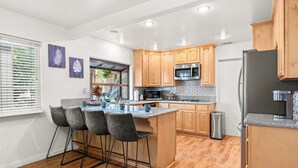 A modern kitchen with wooden cabinets, a central island with bar stools, a stainless steel refrigerator, and a window with blinds.