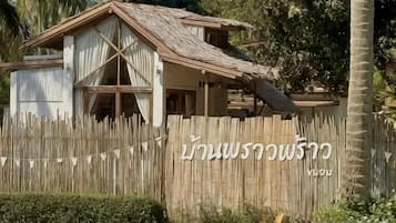 A wooden house with a sloped roof, surrounded by a bamboo fence, and a sign with Thai script.