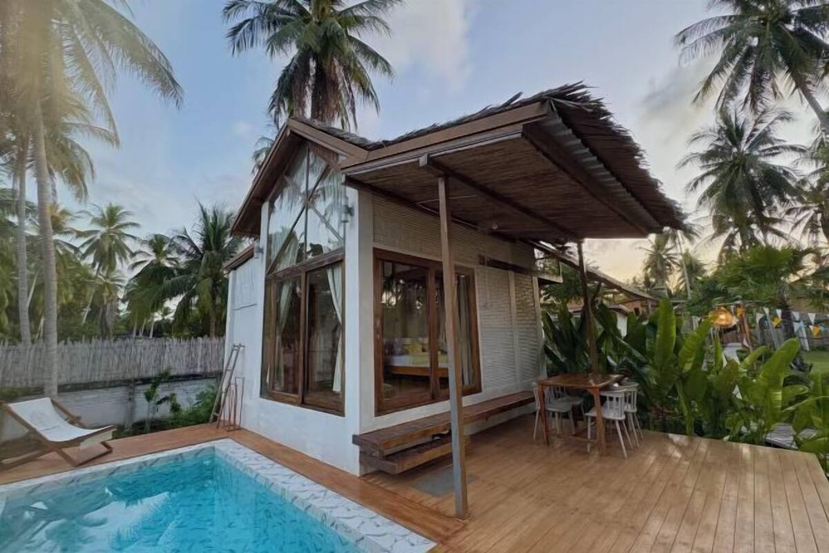 A poolside villa with a wooden deck, a pool, and a view of palm trees.