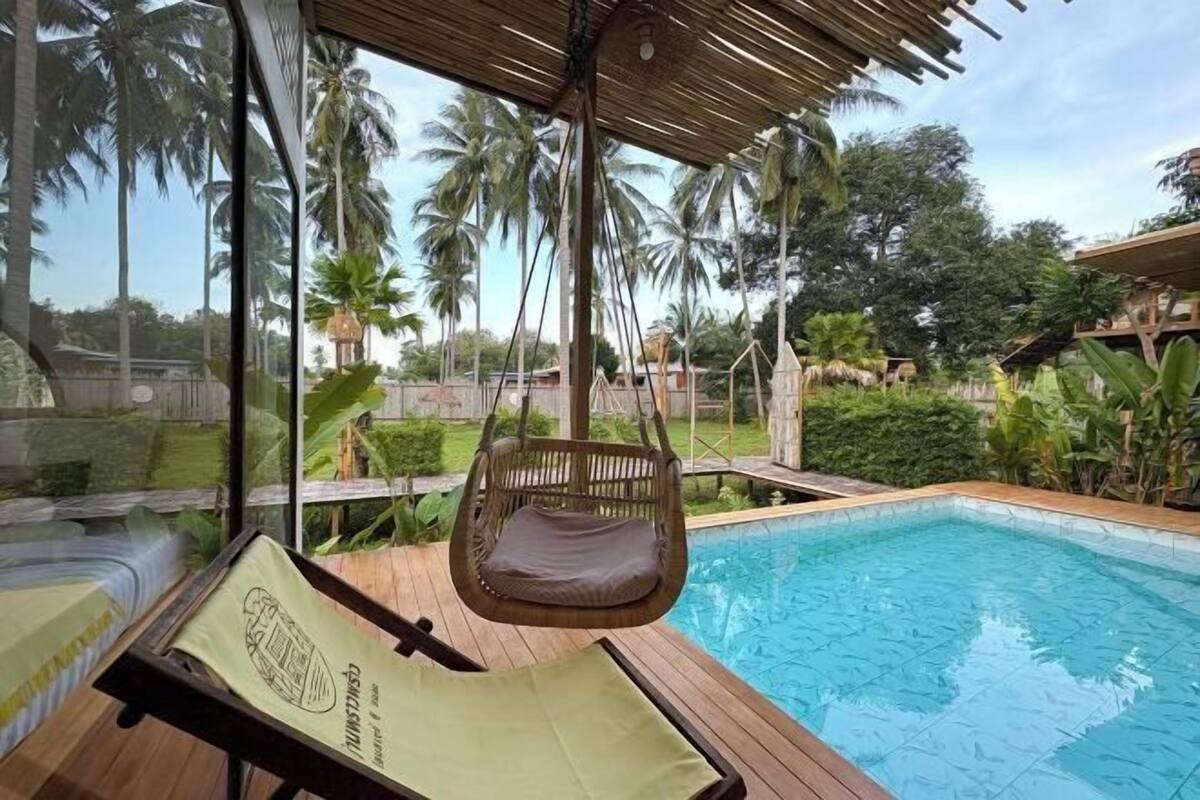 A poolside area with a hanging chair, a lounge chair, and a view of palm trees and a building in the background.
