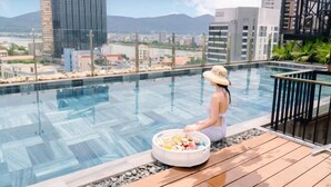 A person sitting by a pool with a cityscape view, a wooden deck, and a basket of snacks. - Tropical Dorm Hostel Da Nang by Haviland (Da Nang)