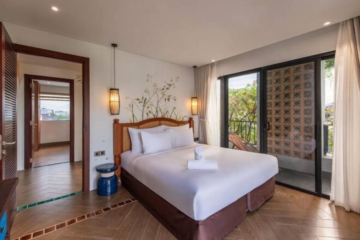 Quarto de hotel com uma cama grande, cabeceira de madeira, mesa de cabeceira, abajur e vista para o exterior por uma porta de correr de vidro.