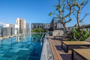 A rooftop swimming pool with a wooden deck, a glass balustrade, and a view of city buildings.
