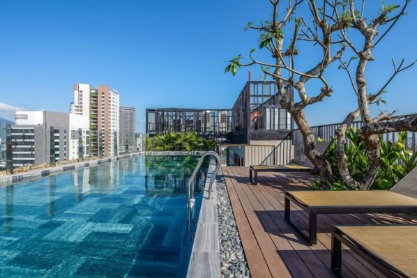 Una piscina en la azotea con terraza de madera, barandilla de cristal y vistas a edificios de la ciudad.