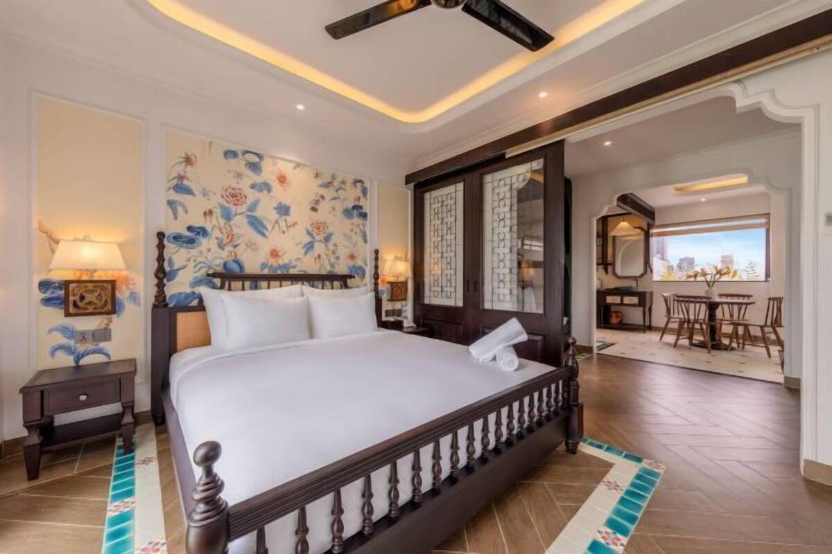 Quarto de hotel com uma cama grande, um ventilador de teto, parede decorativa, uma mesinha com luminária e vista para a sala de jantar.