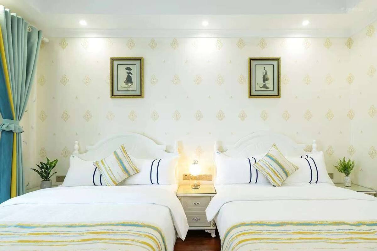 A double room with two beds, a nightstand with a lamp, framed pictures on the wall, and a curtain on the left side.