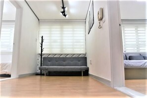 A room with a grey sofa, a window with blinds, a door, and a wall-mounted light fixture. - Seoul Full House/Hongdae Entrance Sta.Exit 7. Party room. Big House. Two Rooms. (Seoul)