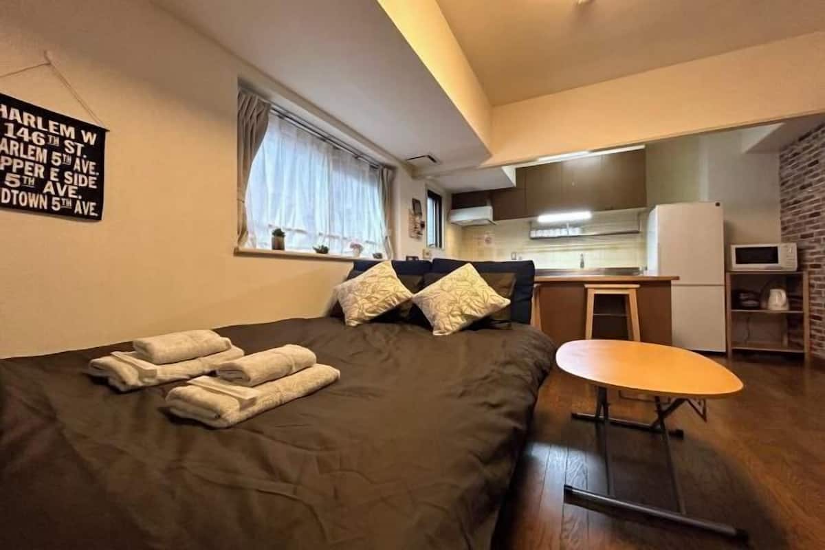 A compact room with a bed, a small table, a kitchenette, and a window with curtains.