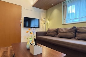 A living room with a brown sofa, a wooden coffee table, a flat-screen TV mounted on the wall, and a window with sheer curtains. - Nestay apartment Tokyo Akihabara (Tokyo)