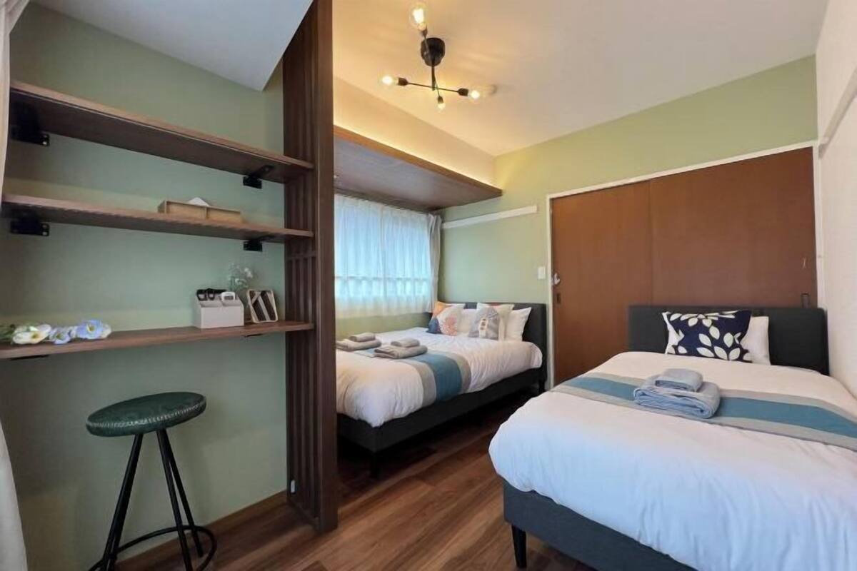 A hotel room with two beds, a wooden wardrobe, a small stool, and shelves with personal items.