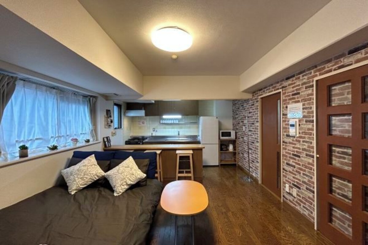Un espacio habitable compacto con kitchenette, un sofá con cojines, una mesita y una ventana con cortinas.