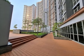 Un edificio residencial moderno con una terraza de madera, un jardín ajardinado y un edificio alto contiguo.