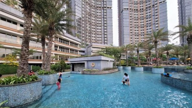 A swimming pool area surrounded by tall residential buildings, palm trees, and a small structure with a circular design. - Gold coast seaview pik Jakarta by SuiteStay (North Jakarta)