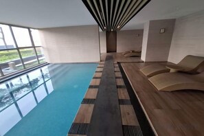 Una piscina con cubierta de madera y sillas reclinables, ventanas amplias y vista a zonas verdes.