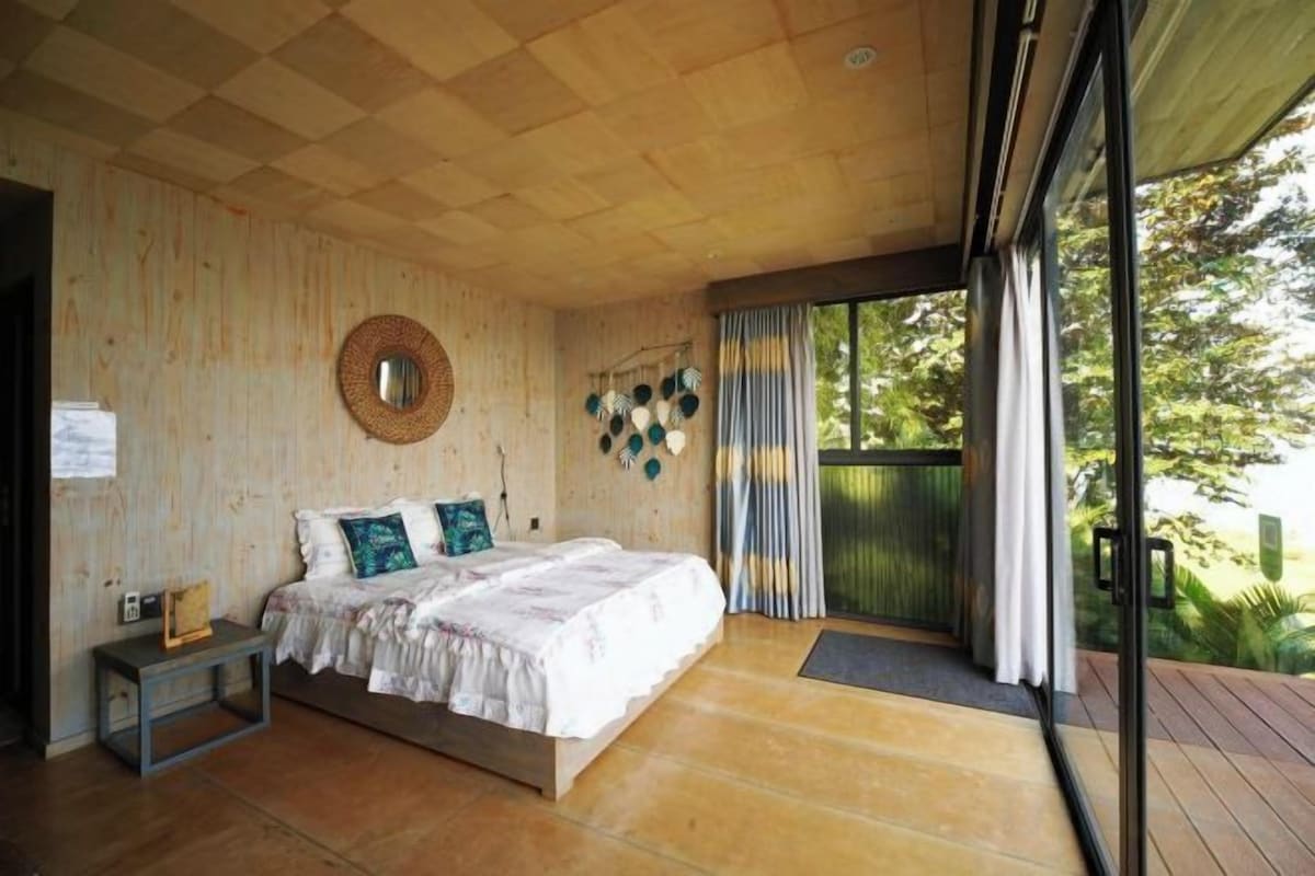 A bedroom with a wooden ceiling, a bed with white bedding, a nightstand, and a view through a glass door to an outdoor area.