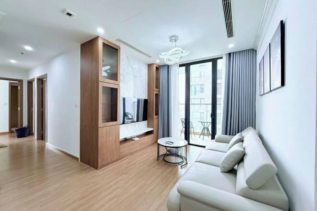 Ein modernes Wohnzimmer mit einer Couch, einem Fernseher und einem kleinen Tisch. Ein großes Fenster mit Vorhängen bietet Blick auf umliegende Gebäude.