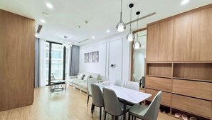 A modern living room with a dining area, a sofa, and a large wooden cabinet with shelves. - PQG - Vinhomes Skylake - Lavish - Resplendent Apartments in Hanoi (Hanoi)