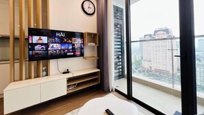 A modern living room with a flat-screen TV mounted on a wooden shelf, a clock on the wall, and a large window offering a city view. - PQG - Vinhomes Skylake - Lavish - Resplendent Apartments in Hanoi (Hanoi)