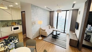 A modern hotel room with a living area, a sofa, a dining table with chairs, a kitchen area, and large windows offering a city view. - PQG - Vinhomes Skylake - Lavish - Resplendent Apartments in Hanoi (Hanoi)
