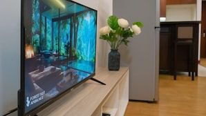 A modern living room with a flat-screen TV displaying a nature scene, a wooden console table, and a vase with white flowers. - The Manor 2 studio- free rooftop pool and free pick up for 5 nights (Ho Chi Minh City)