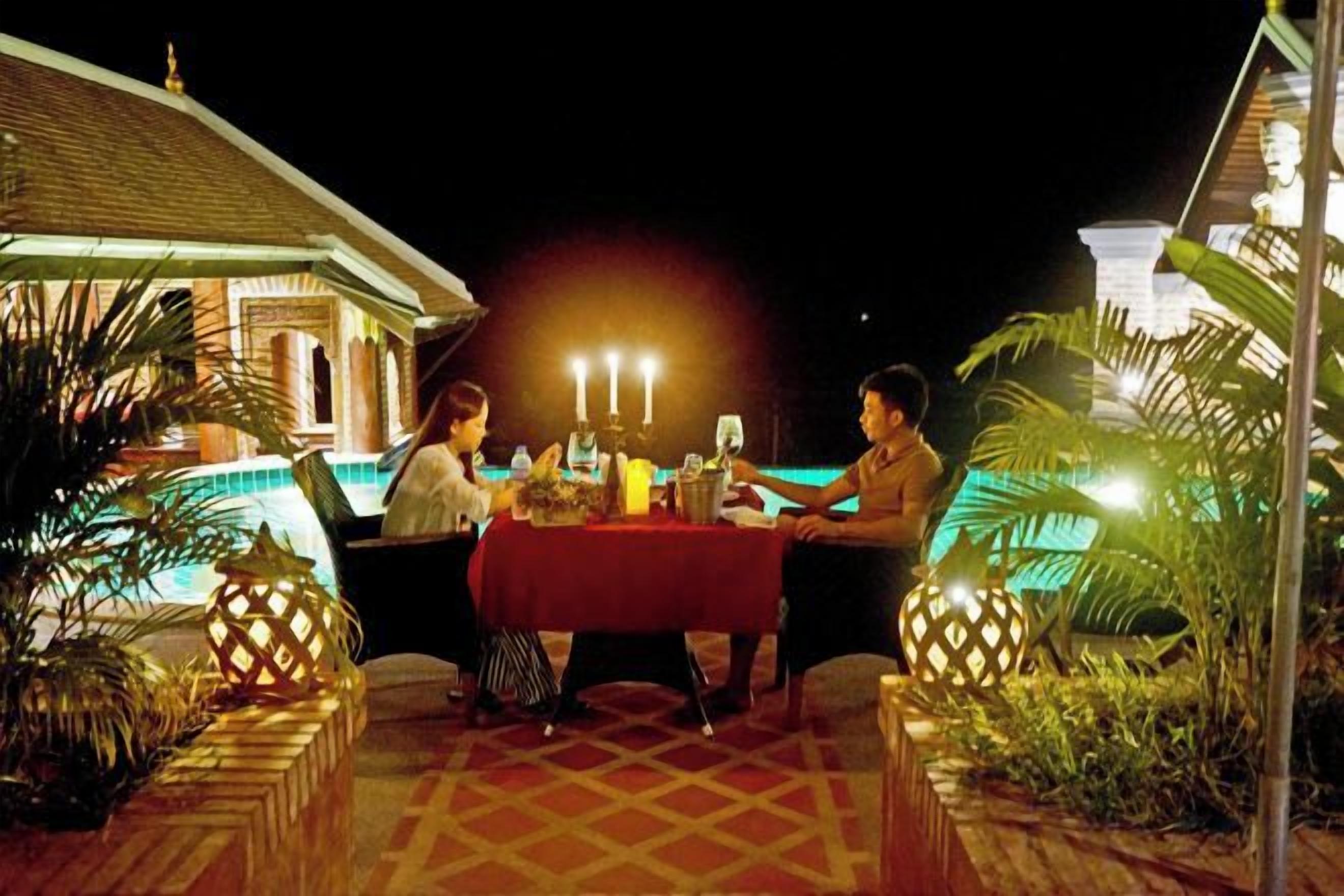 An outdoor dining area with a table set for three, surrounded by plants and a pool in the background.