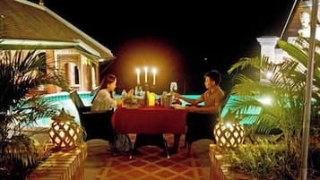 An outdoor dining area with a table set for three, surrounded by plants and a pool in the background.