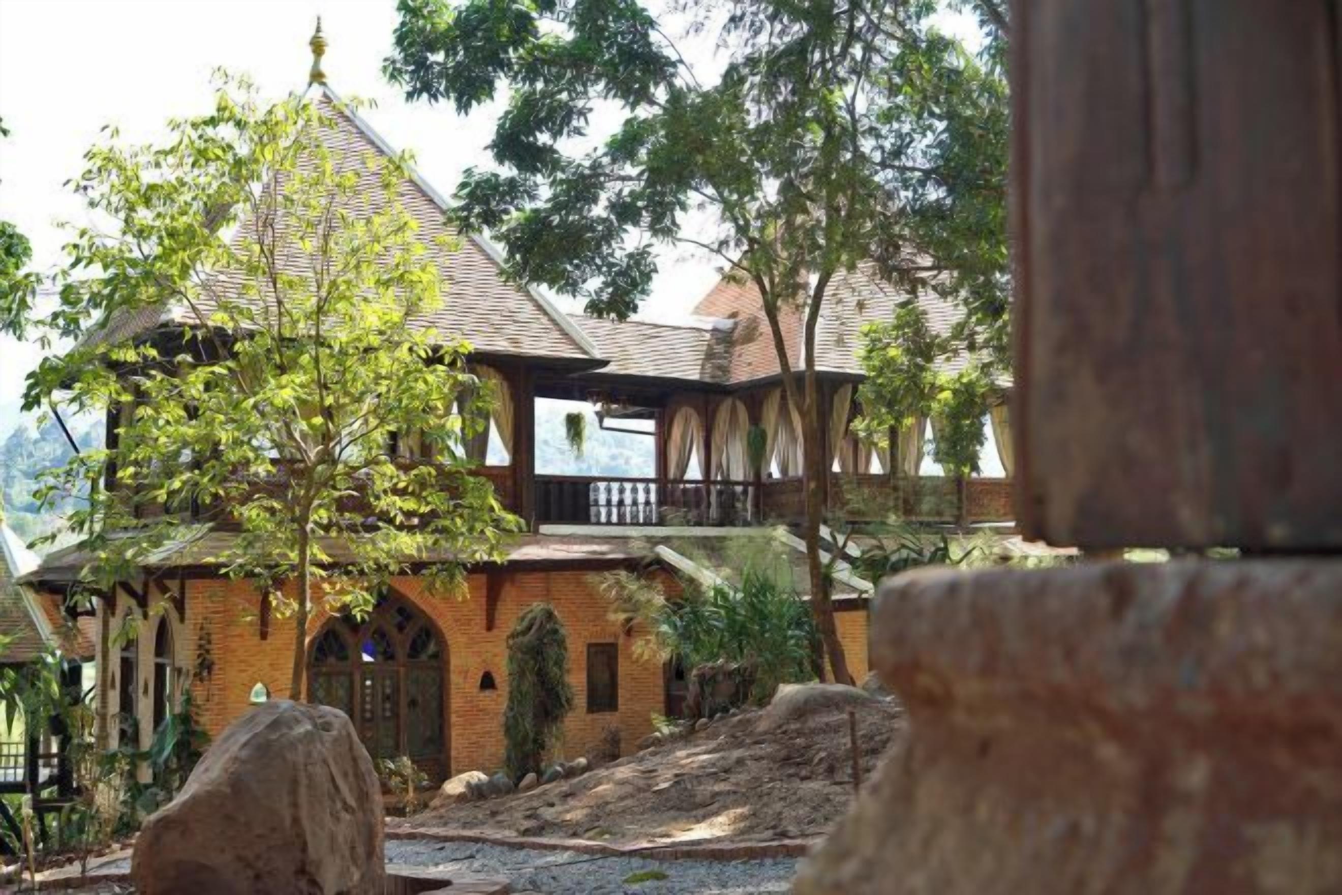 Um edifício de tijolos tradicional com varanda, cercado por árvores e um caminho de cascalho.