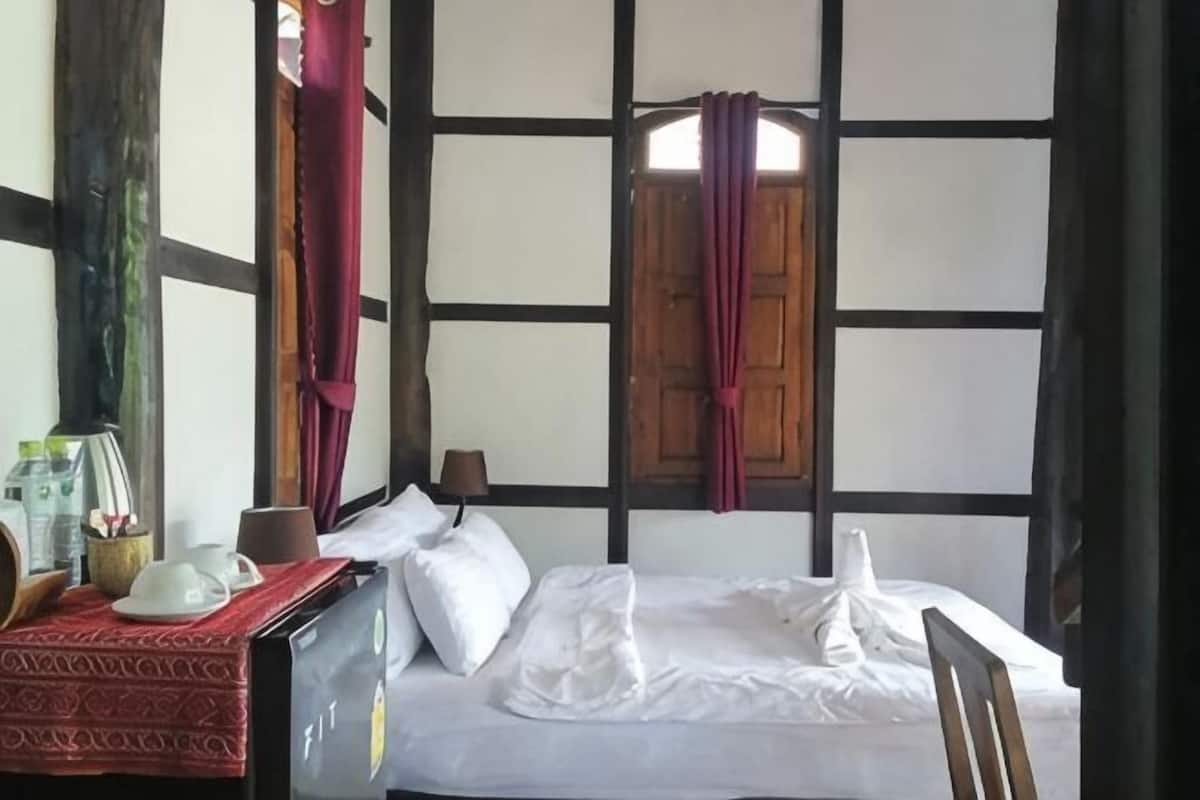 A room with a bed, a table with a red cloth, a window with wooden shutters, and a wall with exposed beams.