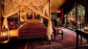 A four-poster bed with a canopy, a wooden chair, a table with a lantern, and a large window with a view of greenery. - Chateau Orientale (Luang Prabang)
