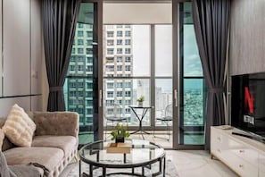 Un salon moderne comprenant un canapé, une table basse en verre, deux fauteuils, un téléviseur et un balcon offrant une vue sur les immeubles de grande hauteur.