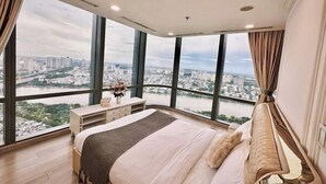 A bedroom with a large bed, a nightstand, and a dresser. There are large windows offering a view of a cityscape and a river. - Condotel Imperial Landmark 81 (Ho Chi Minh City)