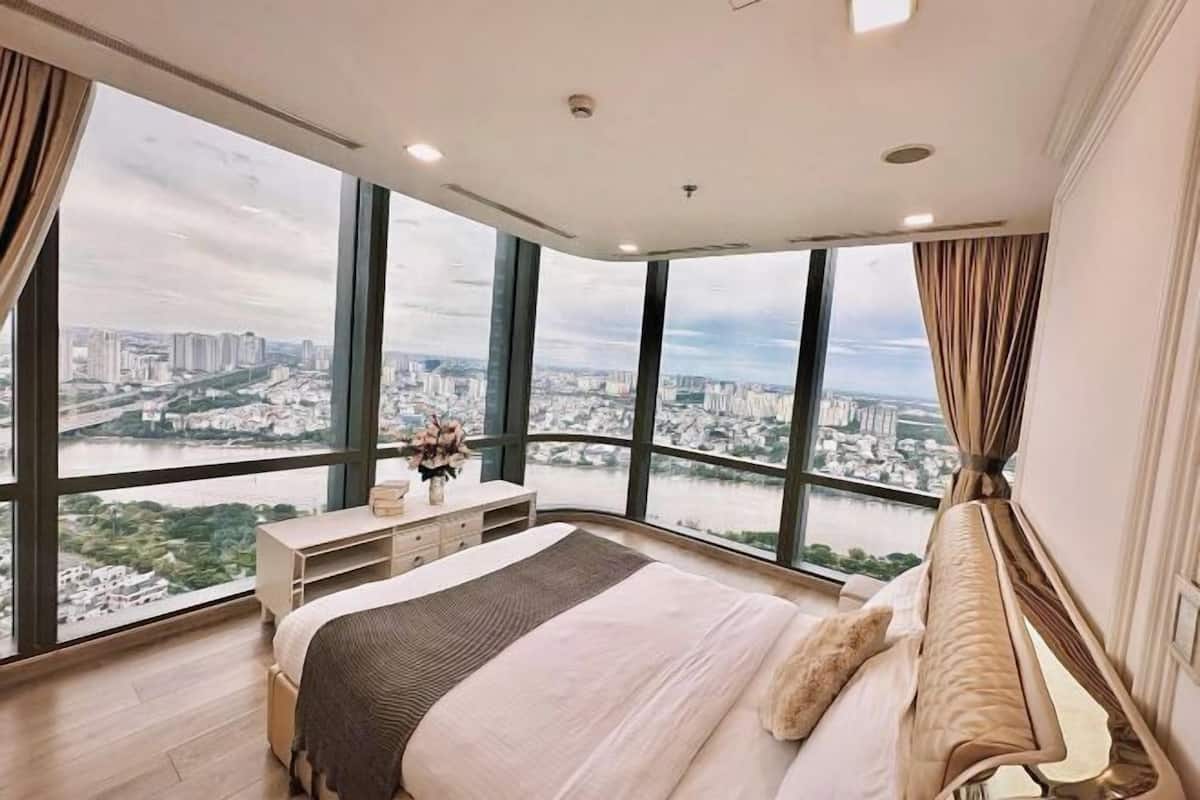 A bedroom with a large bed, a nightstand, and a dresser. There are large windows offering a view of a cityscape and a river.