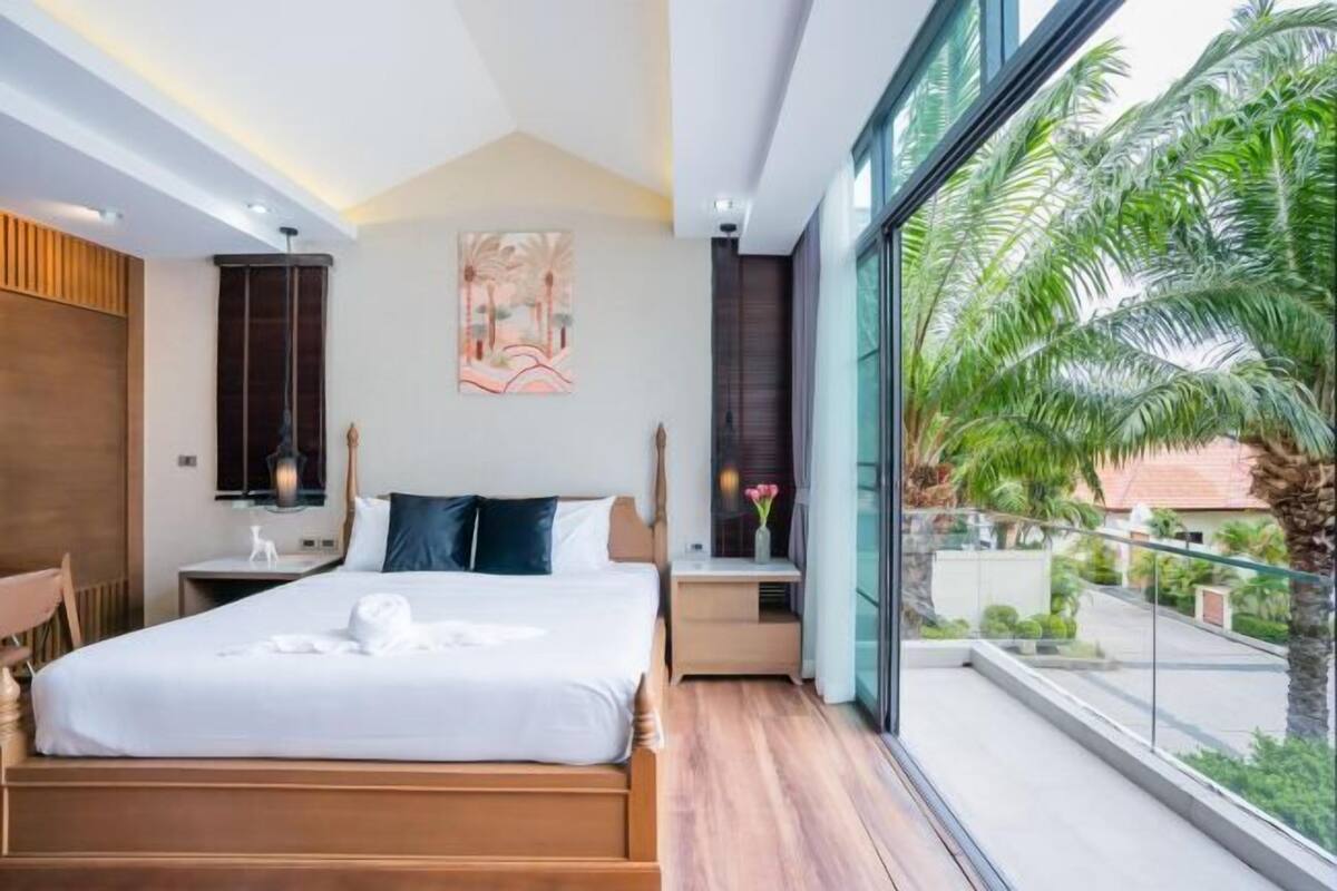 Una habitación de hotel moderna con una cama grande, una mesita de noche, una ventana que ofrece vistas a palmeras y un cuadro en la pared.