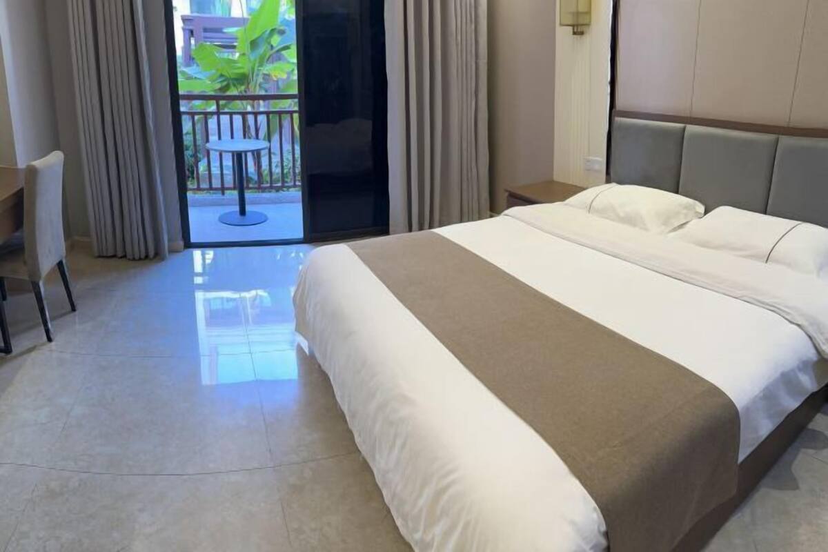 A hotel room with a large bed, a desk, and a balcony with a view of greenery.