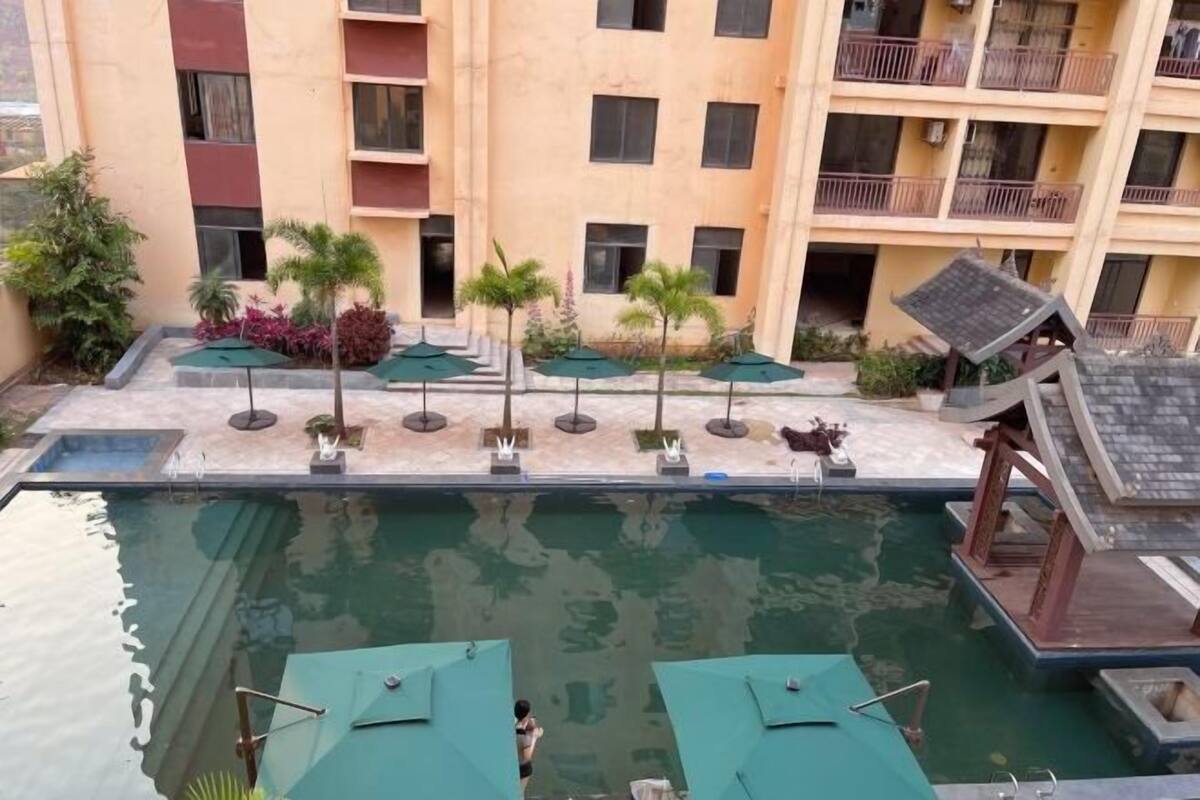 A swimming pool with sun loungers and a small bridge, surrounded by a building with balconies.