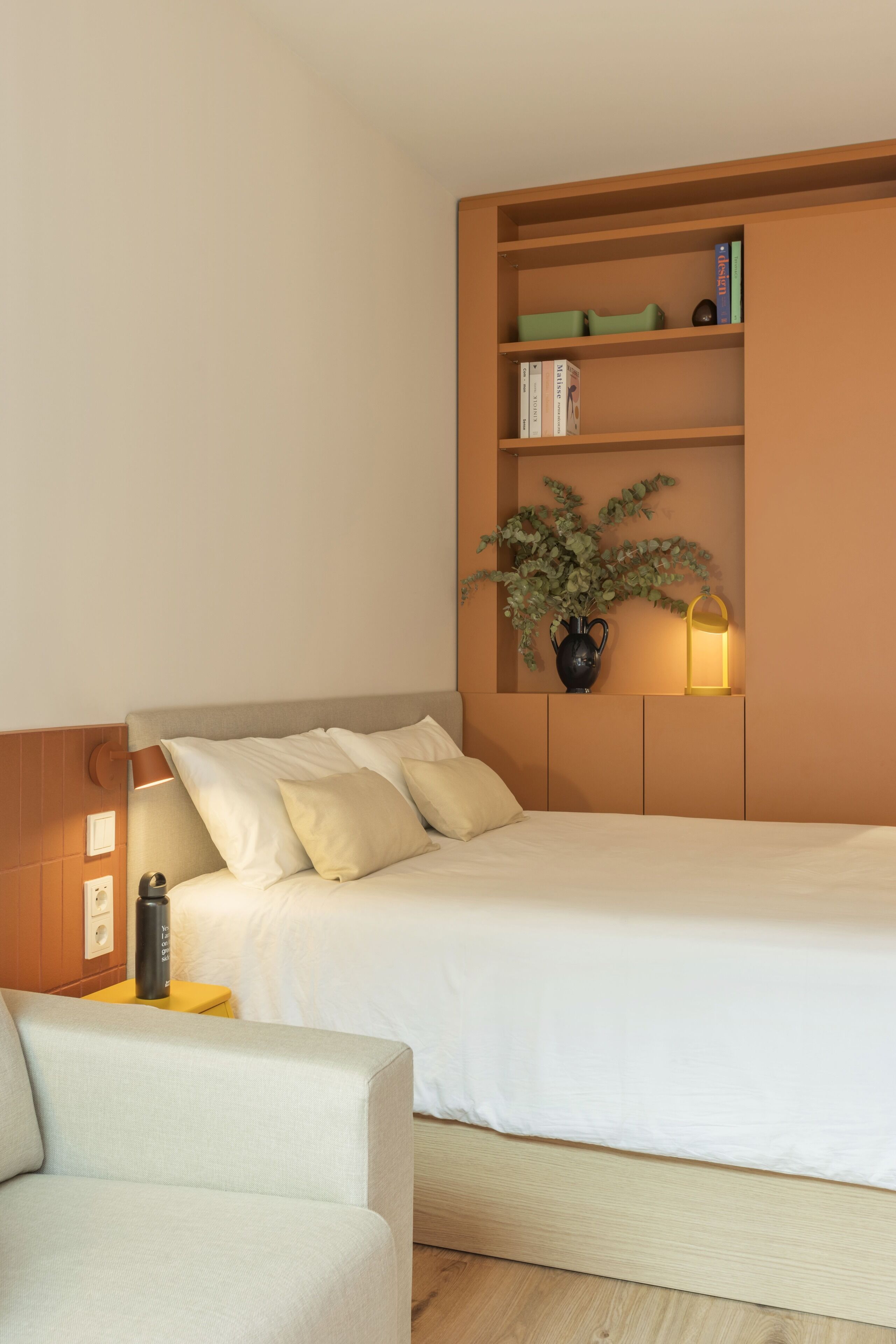 A neatly arranged bedroom with a bed, bedside table, lamp, and a small shelf with books and a plant.