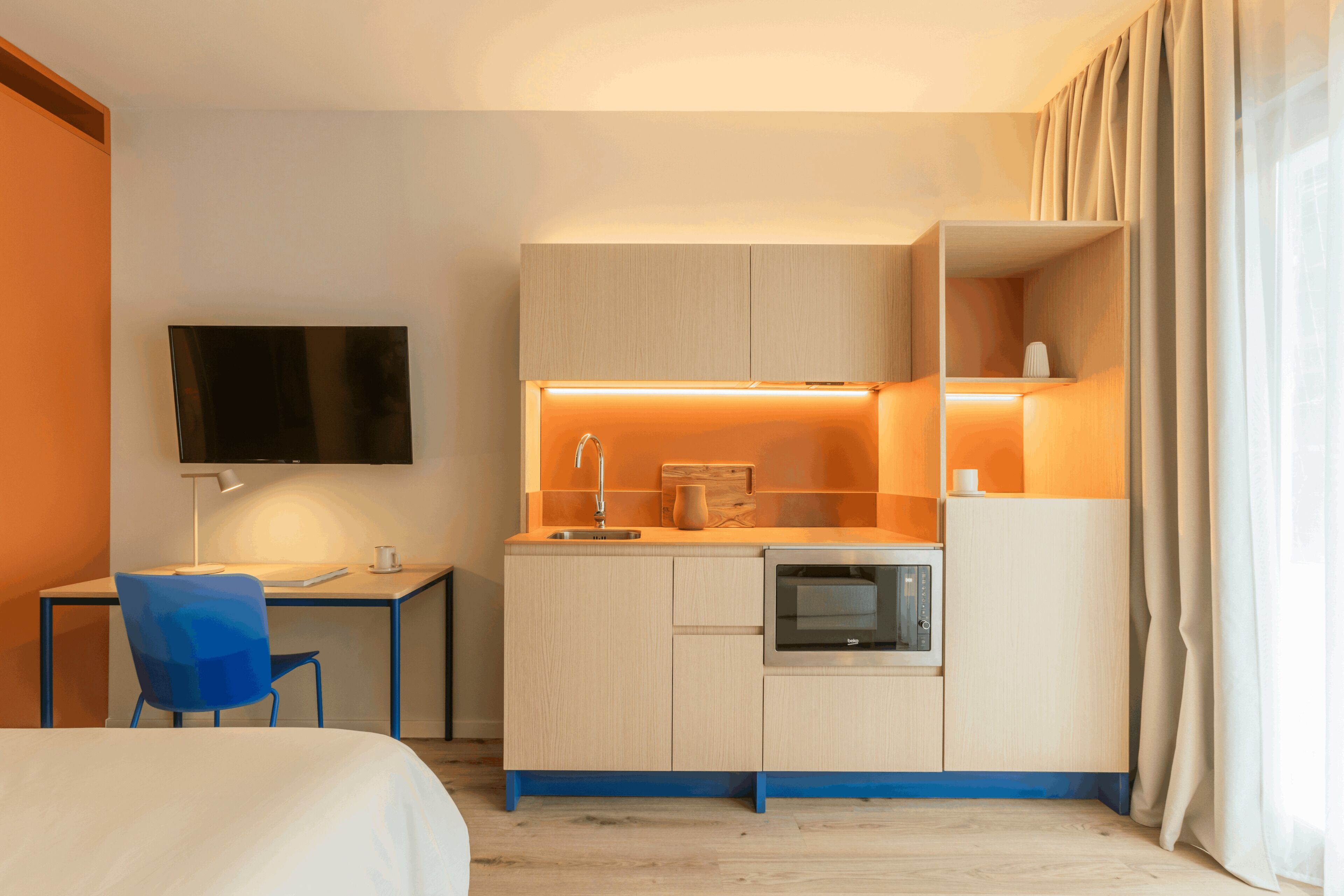 Ein kleines Hotelzimmer mit Küchenzeile, Schreibtisch, Stuhl, Bett und Wandfernseher.