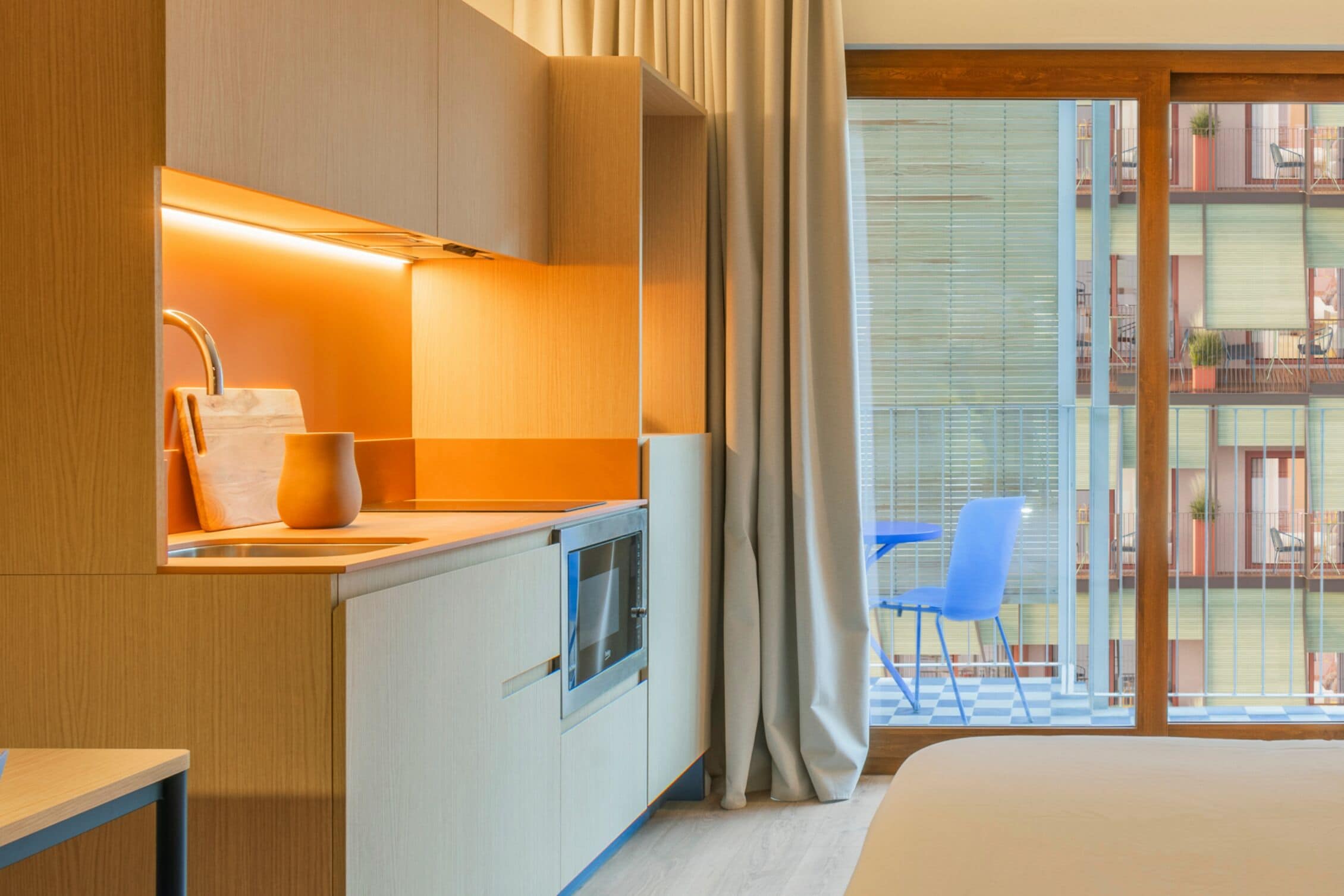 A modern hotel room with a kitchenette, a large window with a view of the balcony, and a bed.