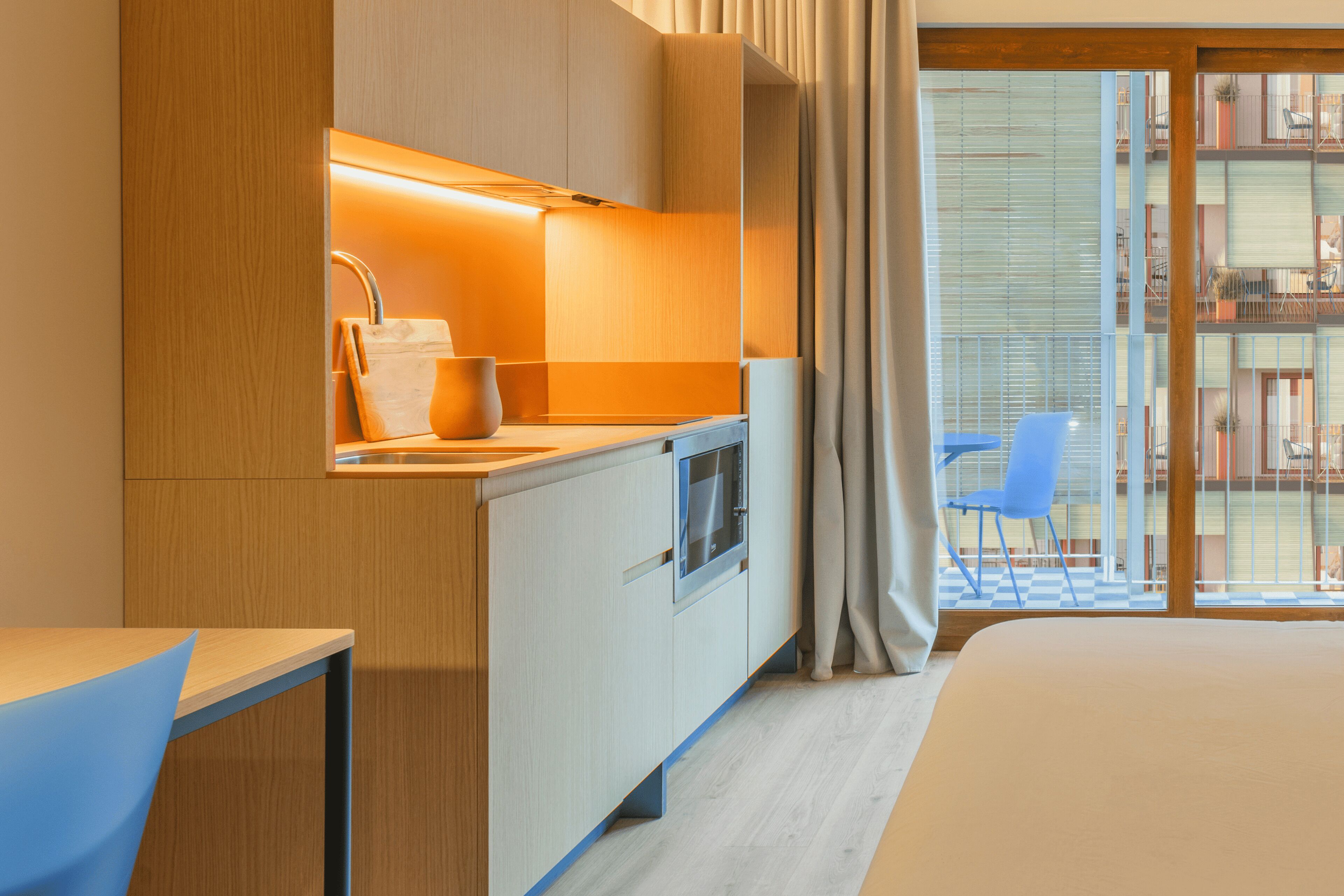 A modern hotel room with a kitchenette, a large bed, a blue chair, and a view of the outside balcony.