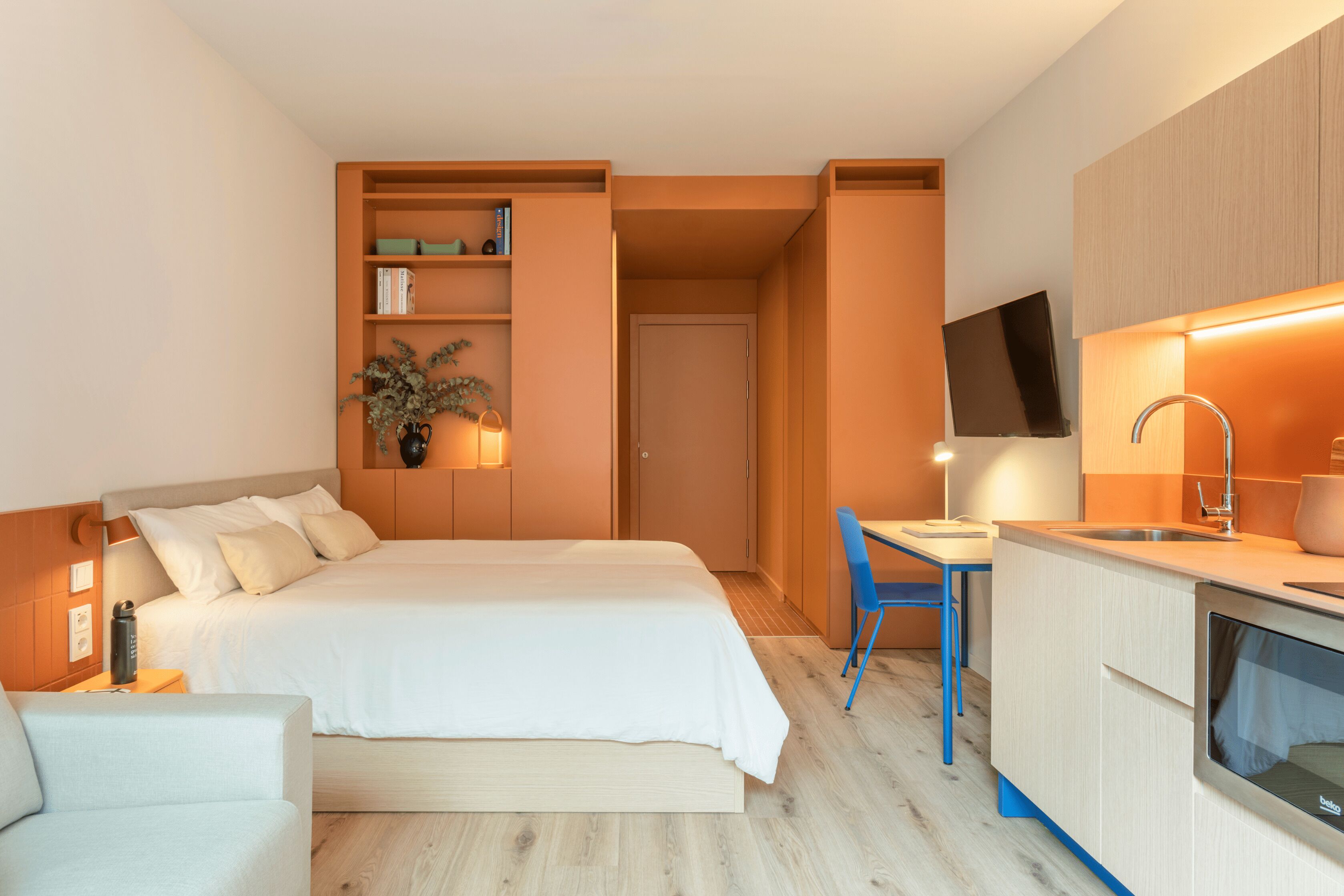 A modern hotel room with a bed, a desk, a chair, a TV, and a kitchenette.