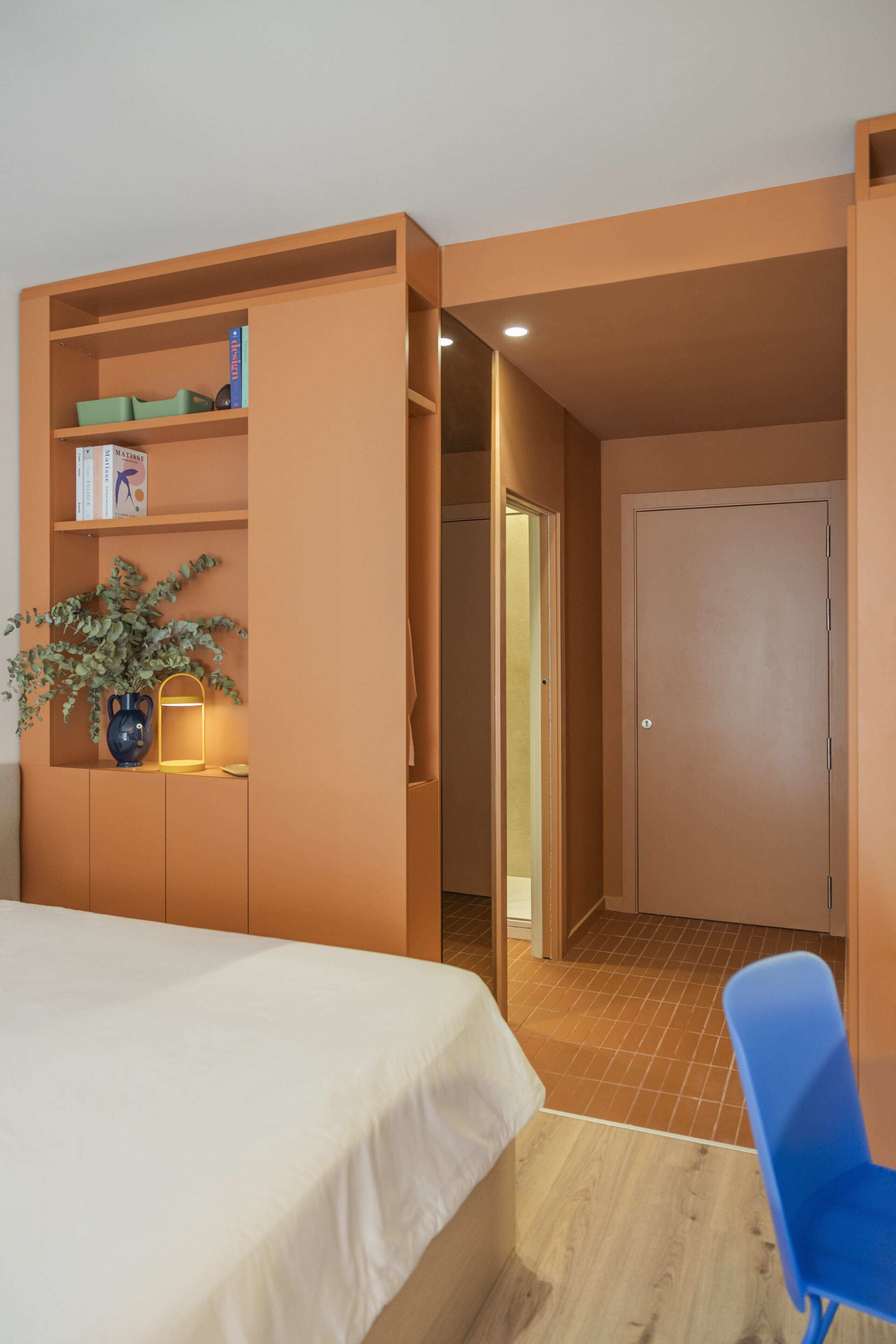 Un dormitorio con cama, una silla azul, una estantería con libros y una planta, y una puerta que da a otra habitación.