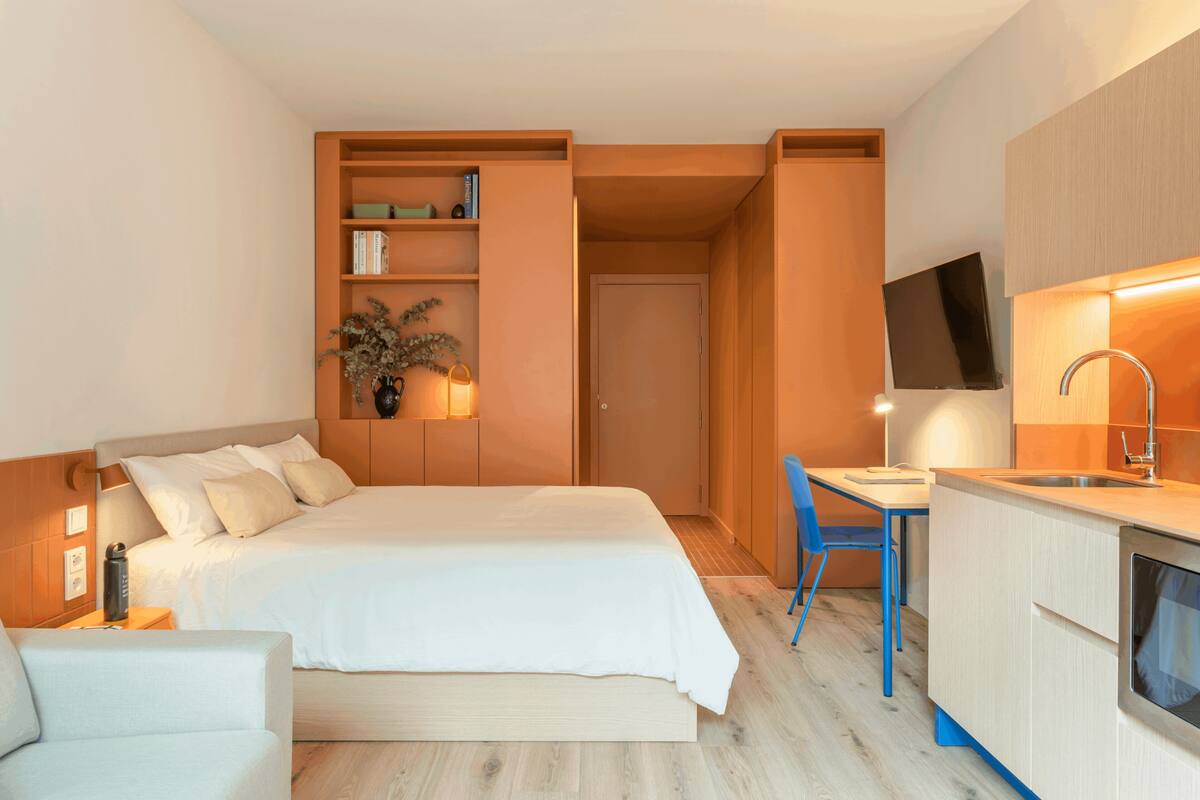 A modern hotel room with a bed, bedside table, chair, desk, and kitchenette.