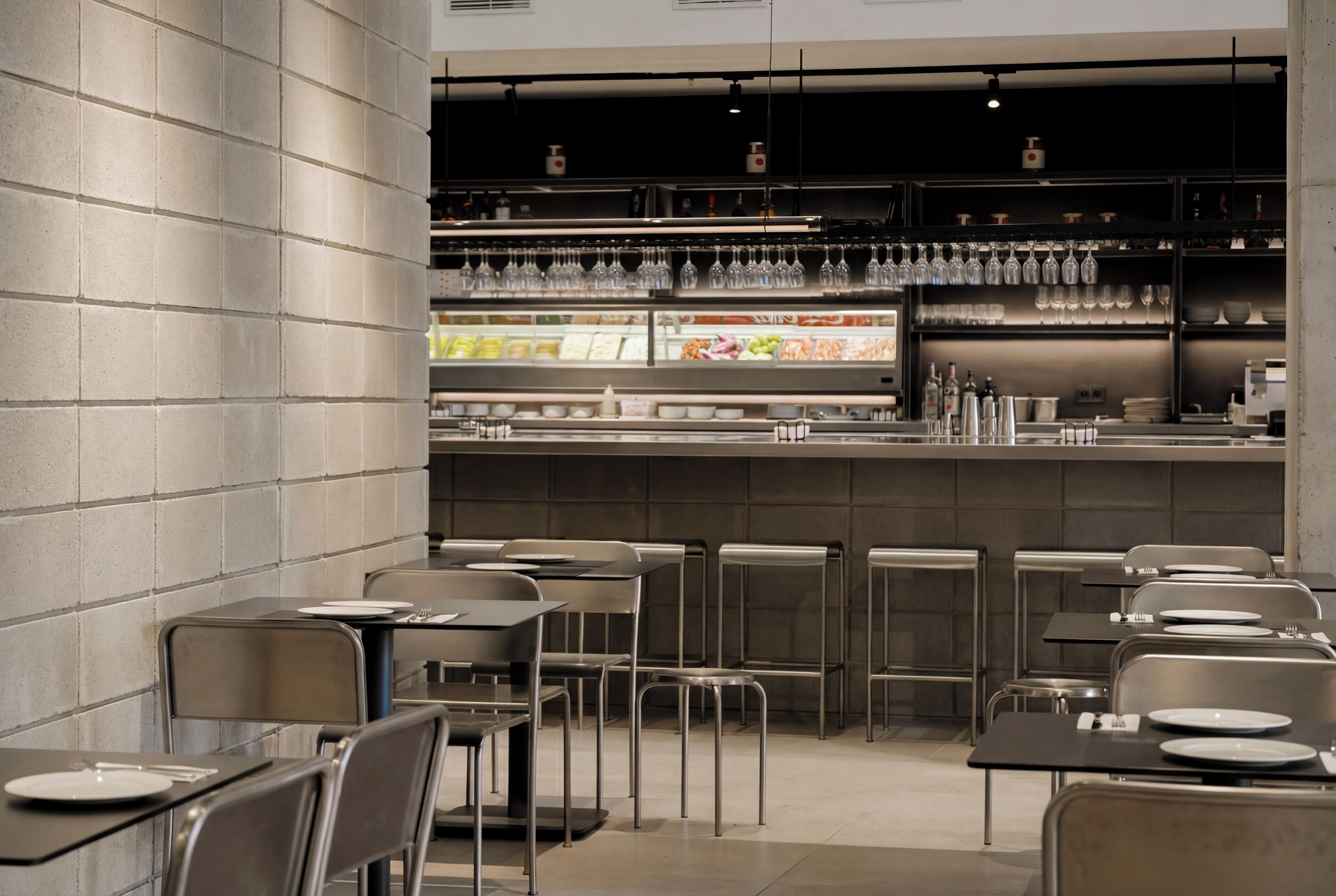 En modern restauranginteriör med metallstolar, bord och en bardisk med hyllor fulla av glas och flaskor.