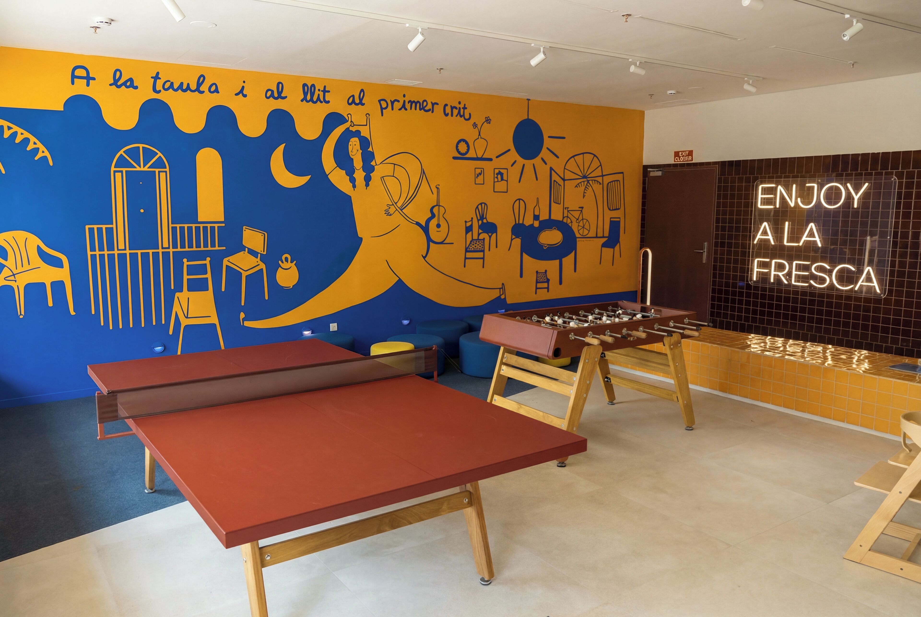A room with a foosball table, a ping pong table, and a mural with text in a foreign language.