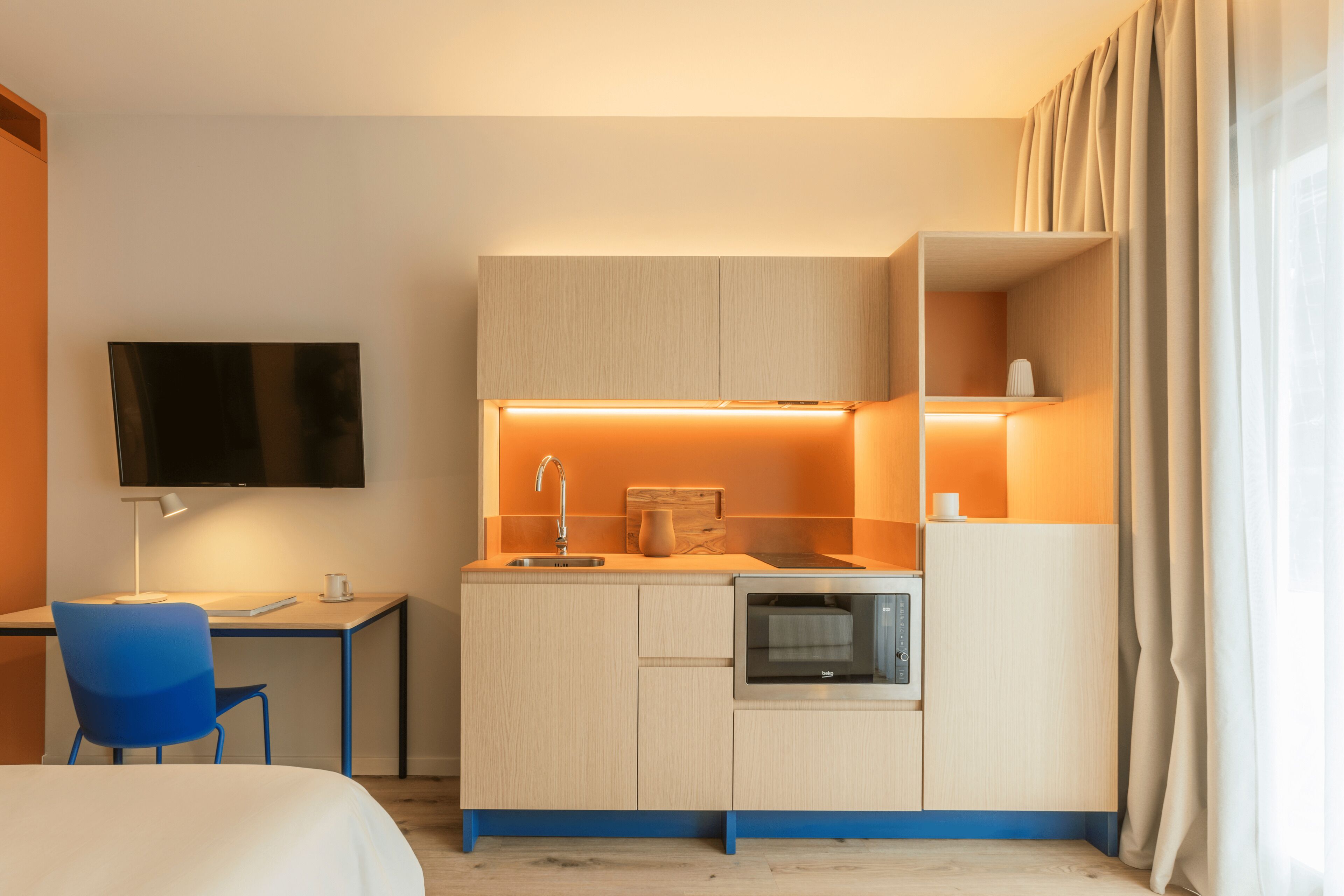 Habitación de hotel compacta con kitchenette, una cama, un escritorio con silla y un televisor montado en la pared.