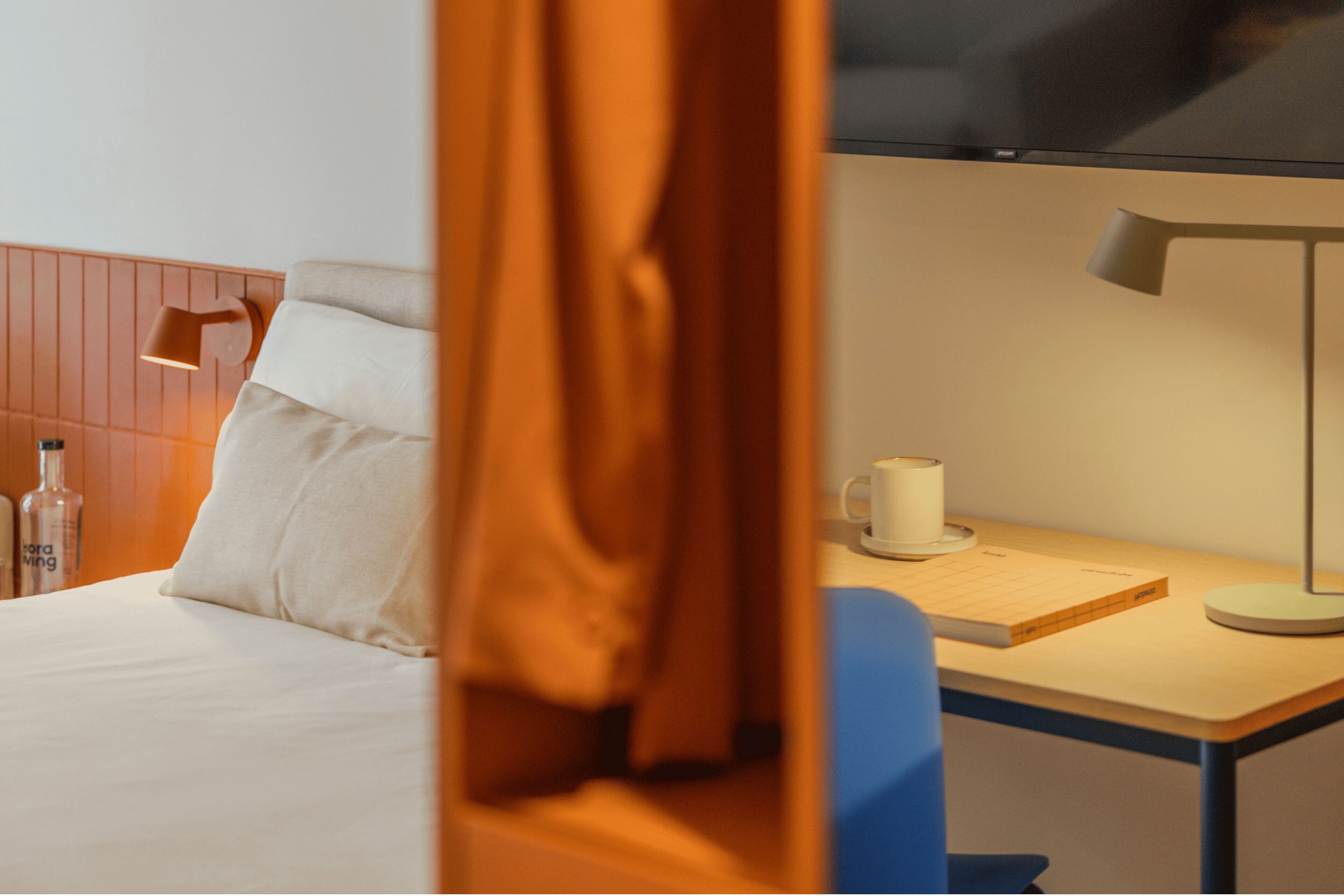 Una habitación de hotel con una cama, un escritorio con lámpara, una taza y un libro.
