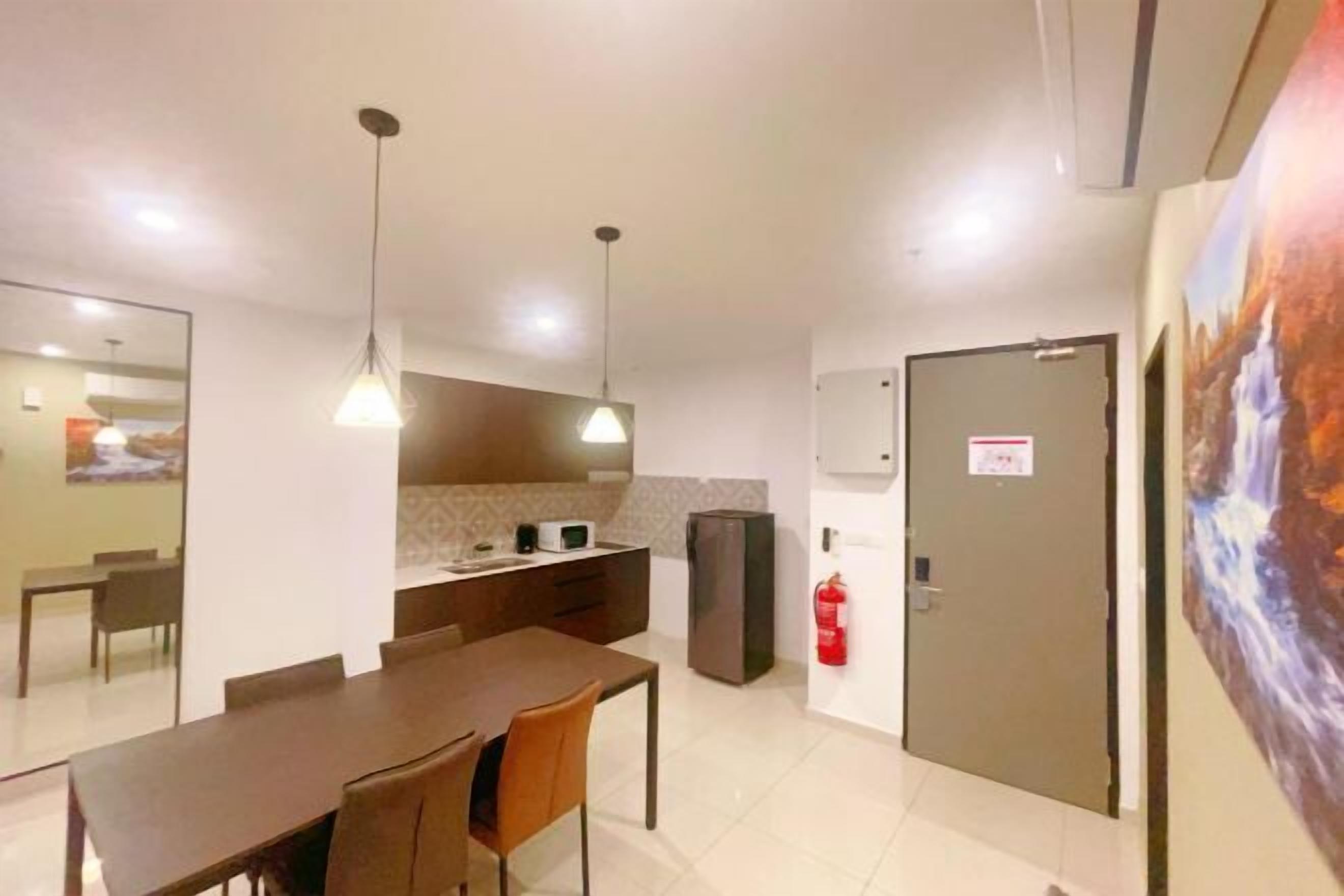 A modern dining area with a long table and chairs, a kitchen area with a refrigerator, and a fire extinguisher mounted on the wall.