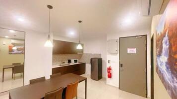 A modern dining area with a long table and chairs, a kitchen area with a refrigerator, and a fire extinguisher mounted on the wall.