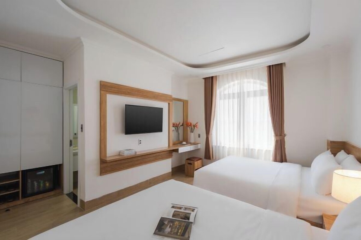 A hotel room with a flat-screen TV mounted on the wall, two beds with white linens, a bedside table with a lamp, and a window with sheer curtains.