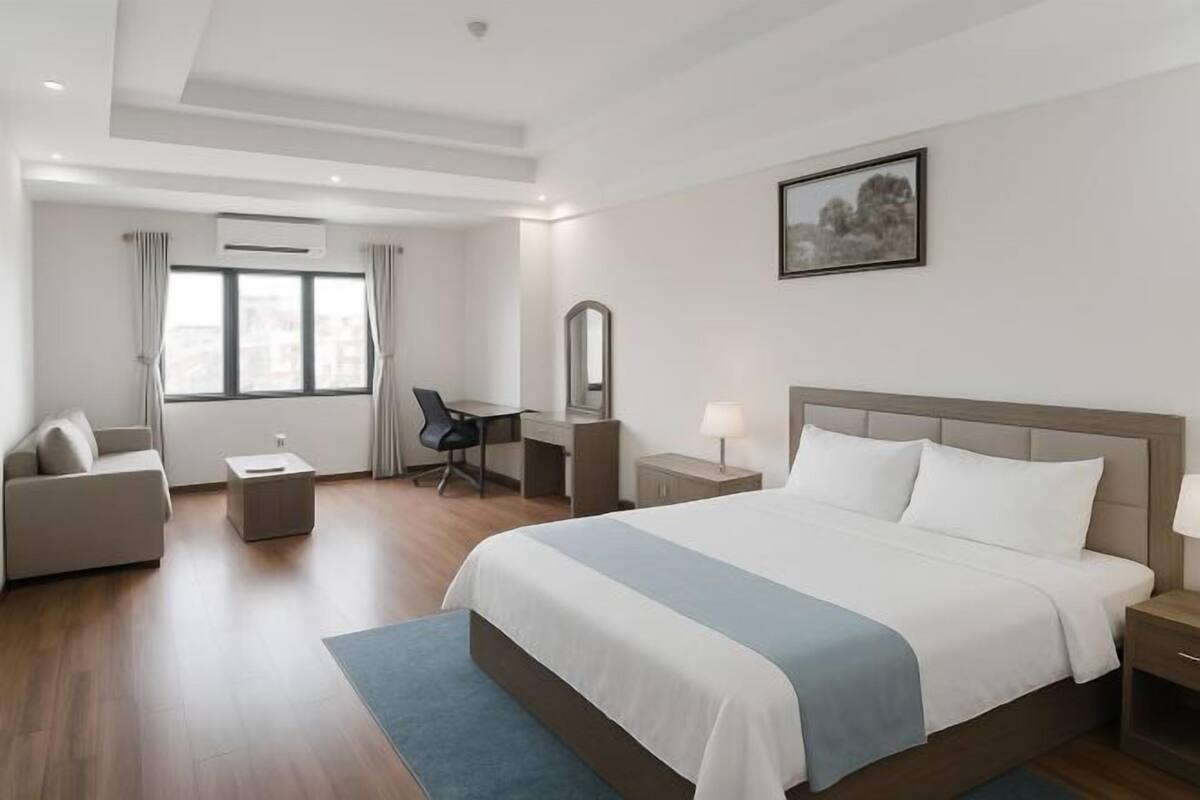 A modern hotel room with a large bed, a desk, a chair, a small table, a nightstand, and a window with curtains.